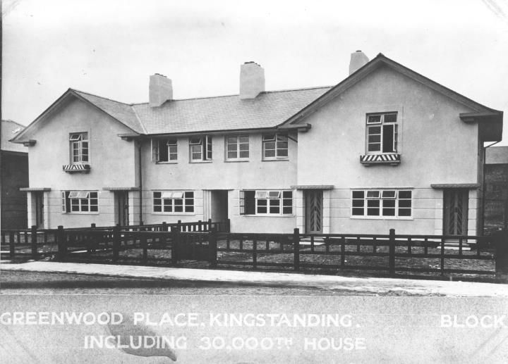 A block of rendered houses with shutters and window boxes behind a timber fence, captioned Greenwood Place, Kingstanding, block including 30,000th house.