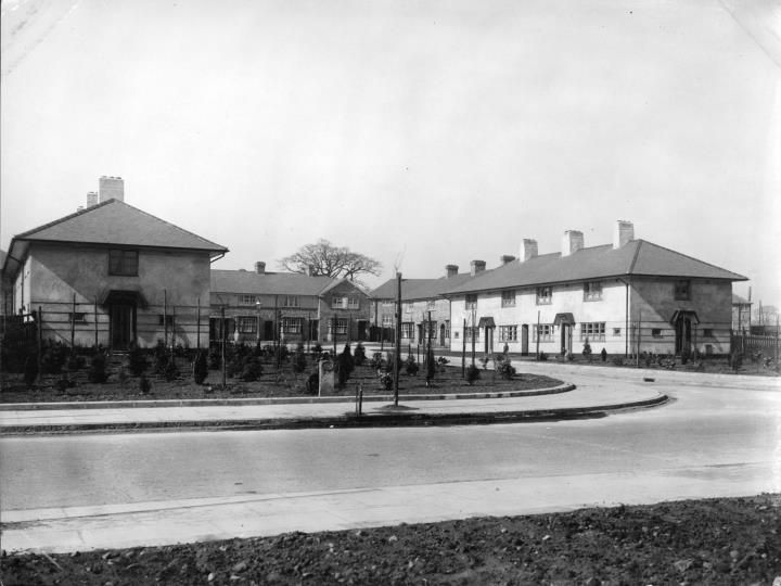 A curving road around a newly planted green, with a single house at the corner and a row of white rendered houses beyond.