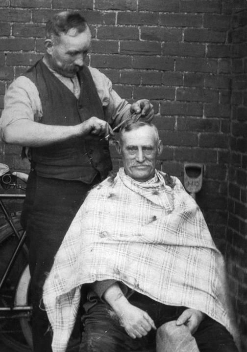 A man in a waistcoat cutting the hair of an older man seated in a checked cape, outside against a brick wall.