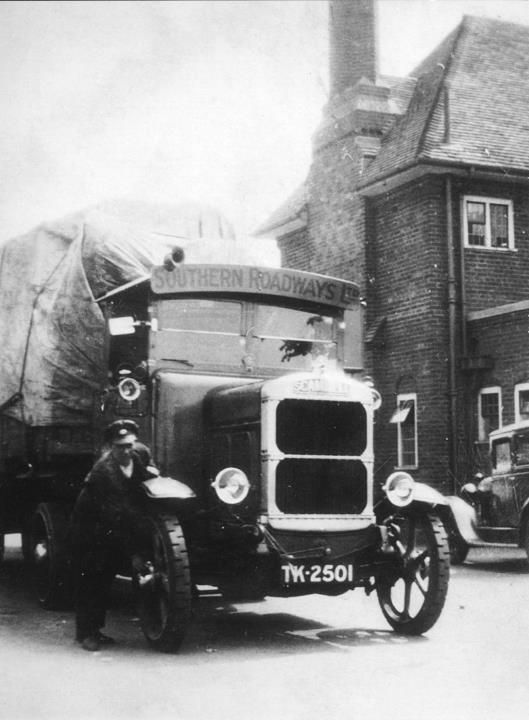 A solid-tyred lorry, registration TK 2501, parked in front of a brick building, with its driver standing beside it.