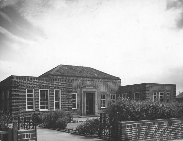 A single-storey brick civic building with a hipped roof and tall windows, behind a low brick wall.