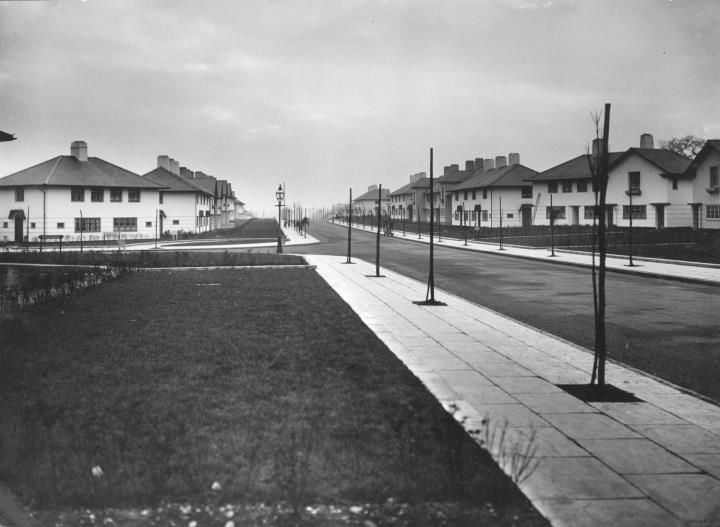 Hurlingham Road looking along its length: wide grass verges, paved footways and newly planted trees on stakes, with white houses on both sides.