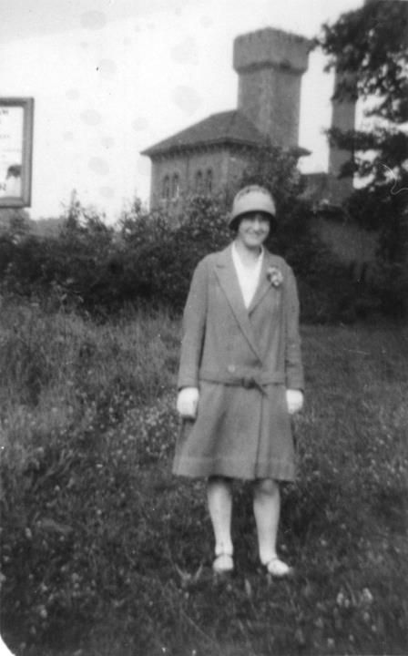 A woman in a 1920s coat and cloche hat standing in long grass, with a towered brick building behind her.