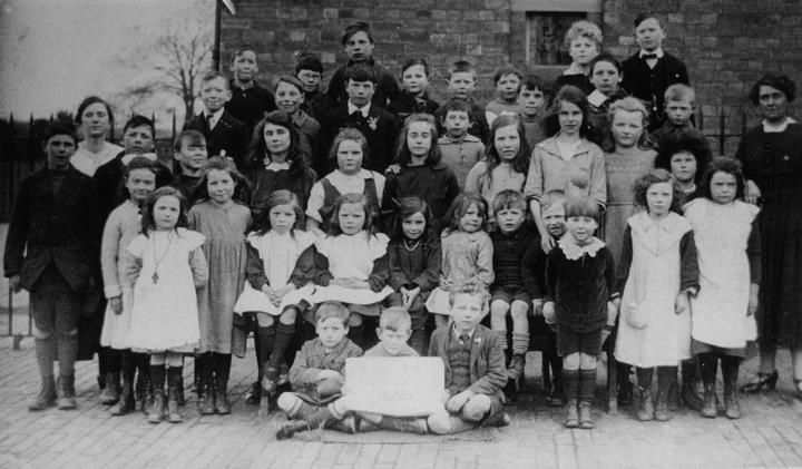 A school class photograph: rows of children with two teachers, a boy in front holding a slate.