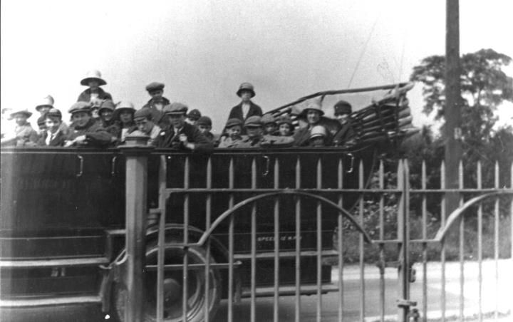 Children in caps and hats crowded into an open charabanc, seen through iron railings and gates.