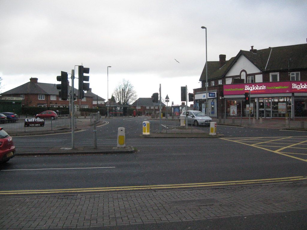 A road junction with traffic lights looking over to Warren Farm Road, with a parade of shops on the right.