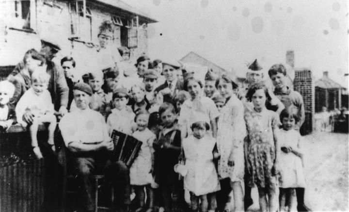 A large crowd of children and adults posed in a street in front of houses, one man holding an accordion.