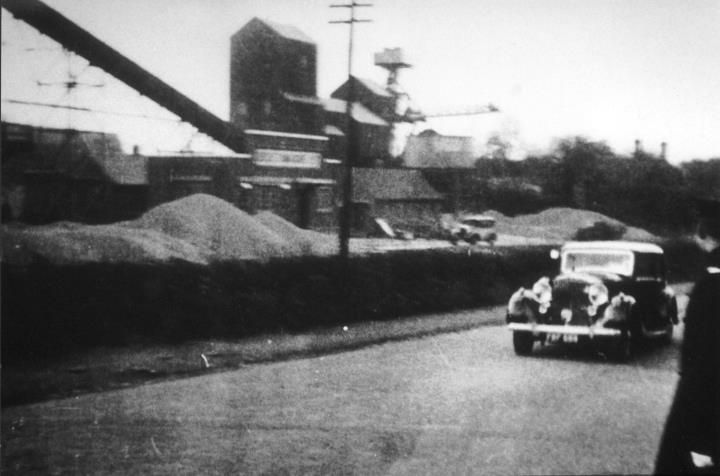 A 1930s car passing an industrial building with a tall hopper and conveyor, beside heaps of gravel.