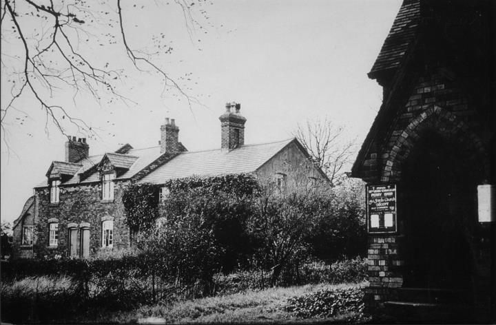 An old brick house with dormer gables behind a garden, with the end of a brick building bearing a notice board in the foreground.