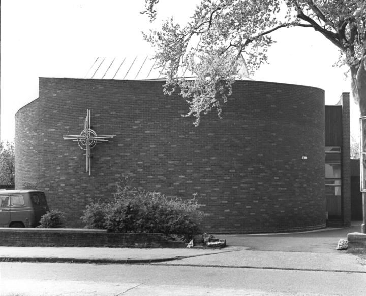 The curved brick wall of St Mark's Church with a metal cross on its face and a van parked alongside.