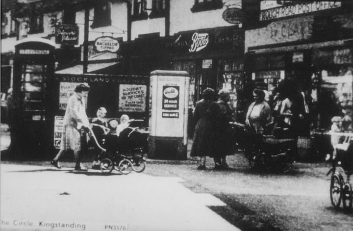 Shoppers and a woman with a pram outside shops at the Circle, beside a telephone box; the print is captioned The Circle, Kingstanding.