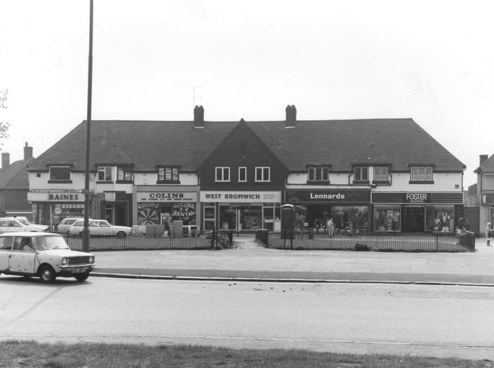 A parade of shops beneath a long tiled roof at the Circle, with shop signs including Baines and Lennards, and a parked car in front.