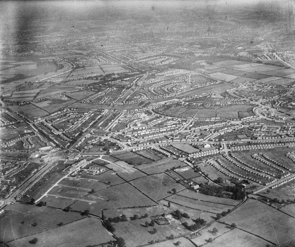 Oblique aerial photograph from the north-west: fields and hedgerows in the foreground, with crescents and rows of new houses beyond.