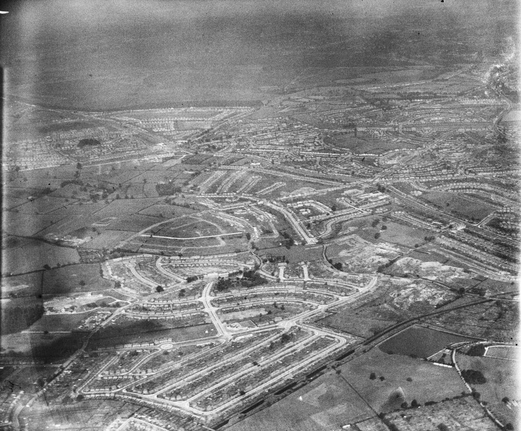 Oblique aerial photograph from the south-west: newly laid-out curving streets of houses in the foreground, some unfinished, with the older estate beyond.
