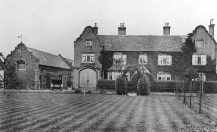 A large brick farmhouse with shaped Dutch gables and a barn, behind a striped lawn.