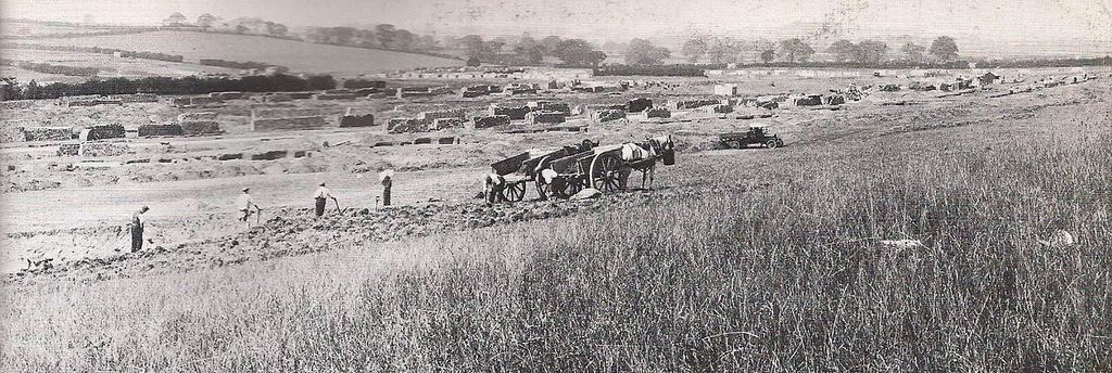 A wide panorama of open field being prepared for building: workmen with hand tools, horse-drawn carts and a lorry, with materials stacked in rows and hedgerow trees on the skyline.
