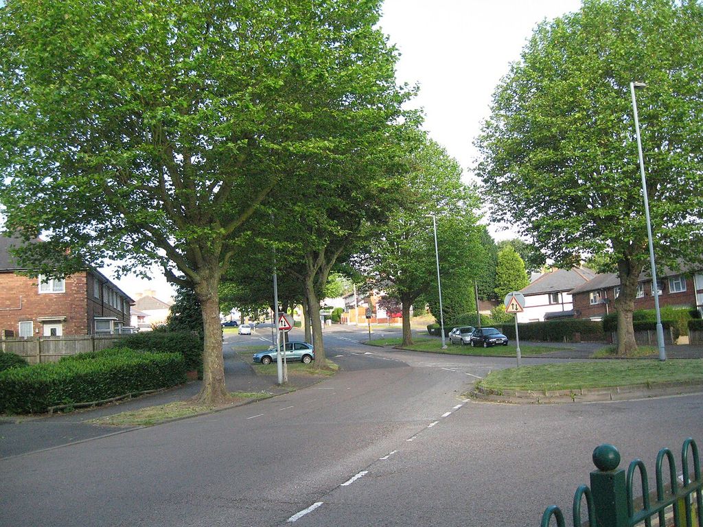 Warren Farm Road curving away beneath mature trees in full leaf, with brick houses on either side.