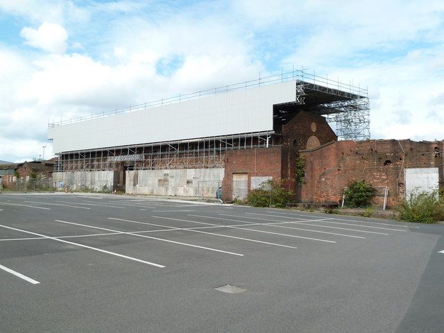 An old brick industrial building partly wrapped in scaffolding and a white temporary cover, behind an empty car park.