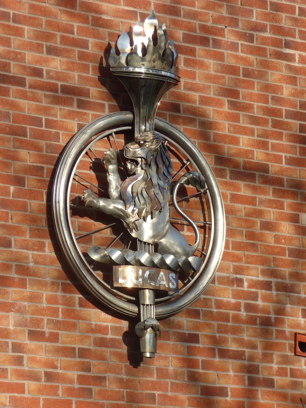 A stainless-steel emblem of a lion standing on a wheel holding a flaming torch, fixed to the brick end wall of a house.