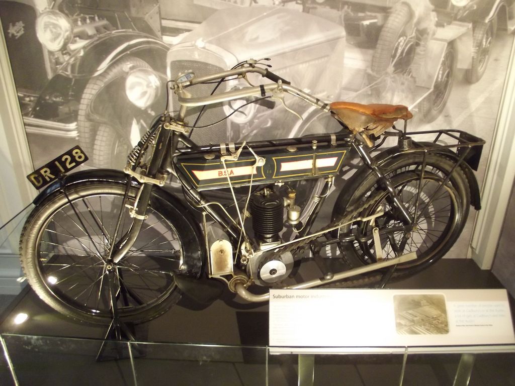 An early black motorcycle with a leather saddle and a single-cylinder engine in a museum display case.