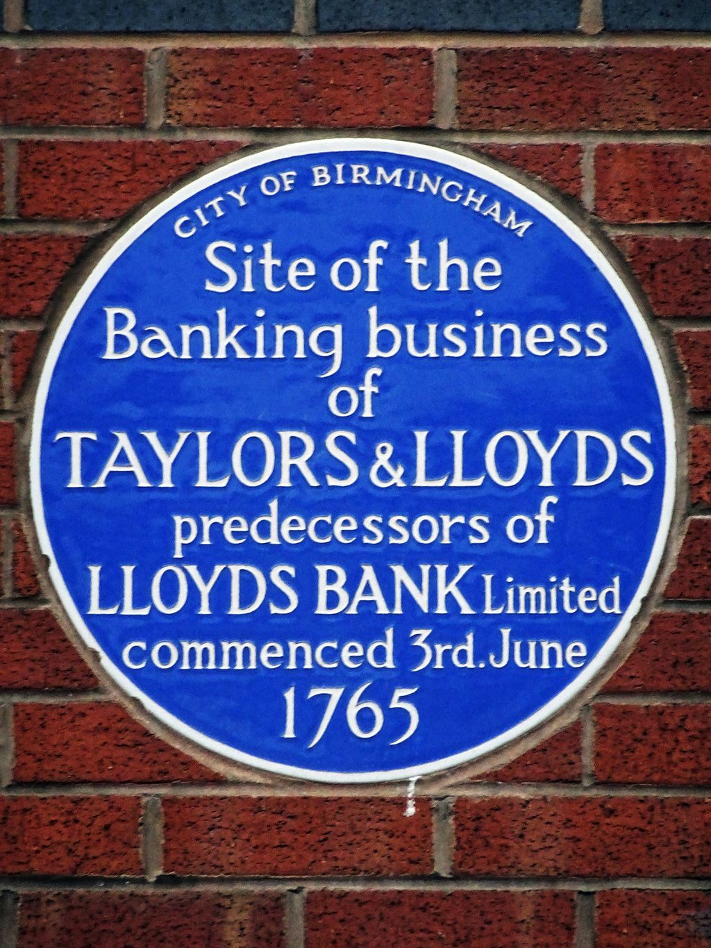 A round blue plaque on a wall, with white lettering about the Taylors & Lloyds bank.
