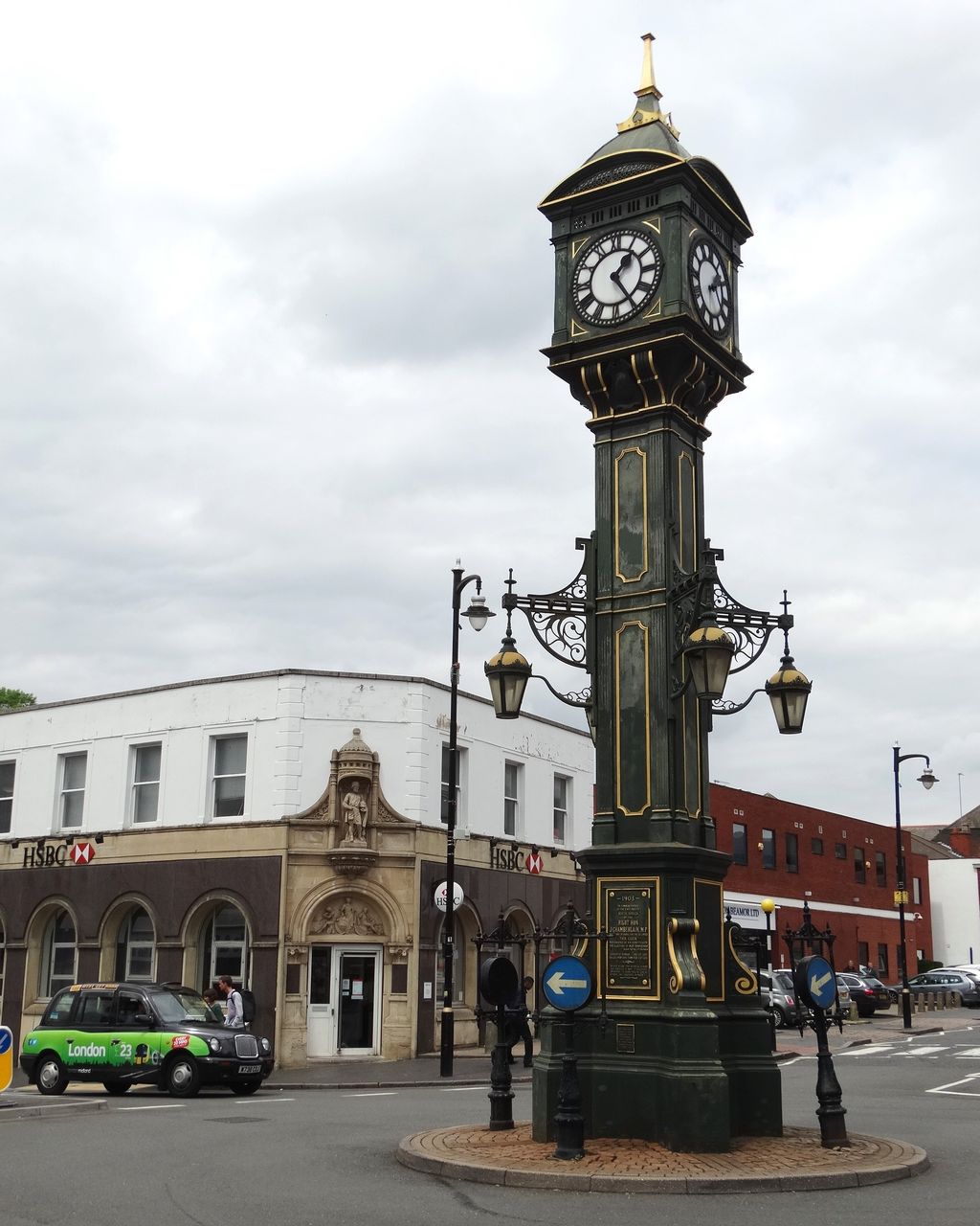 A tall green-and-gold cast-iron clock tower at a road junction, with four clock faces under a small dome.