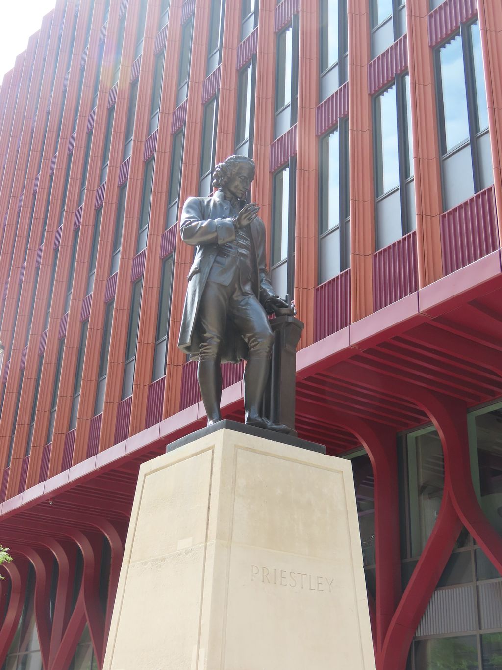 A dark bronze statue of a man in eighteenth-century dress on a tall stone plinth beside a walkway.