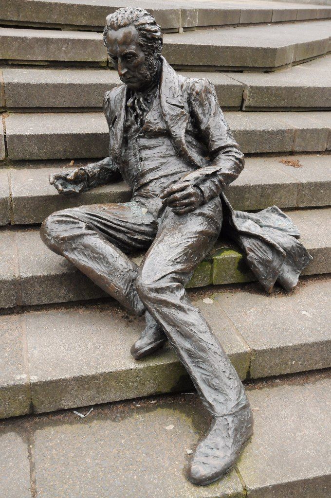 A bronze figure of a man in a long coat sitting on stone steps, a sheet of paper in his hand.