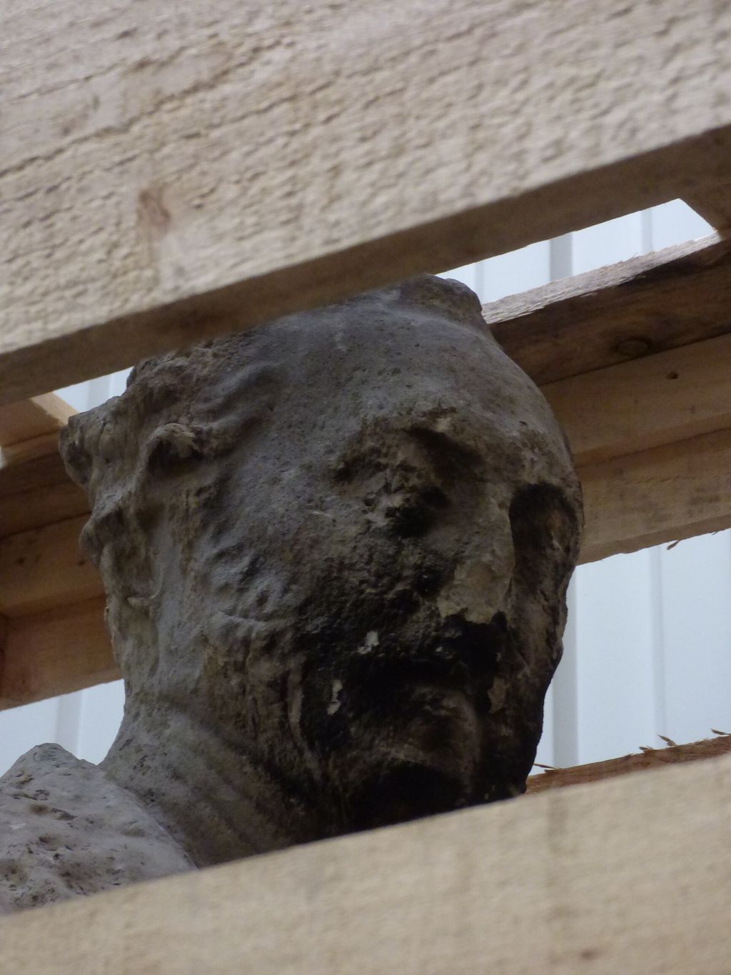 The weathered, damaged marble head of a statue, seen between the slats of a wooden crate.