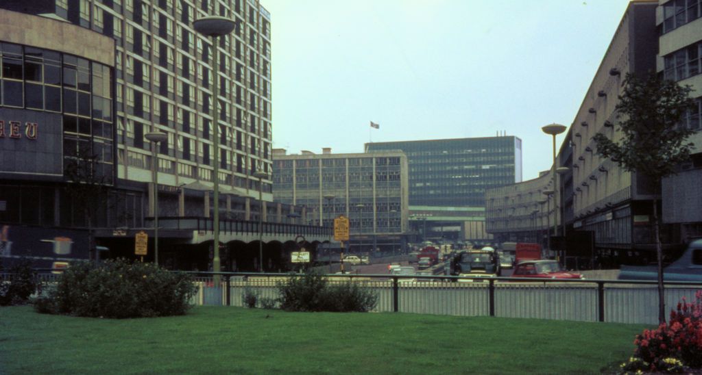 View from a grassy roundabout along a dual carriageway lined with new office blocks and a tall hotel, with traffic in the distance.