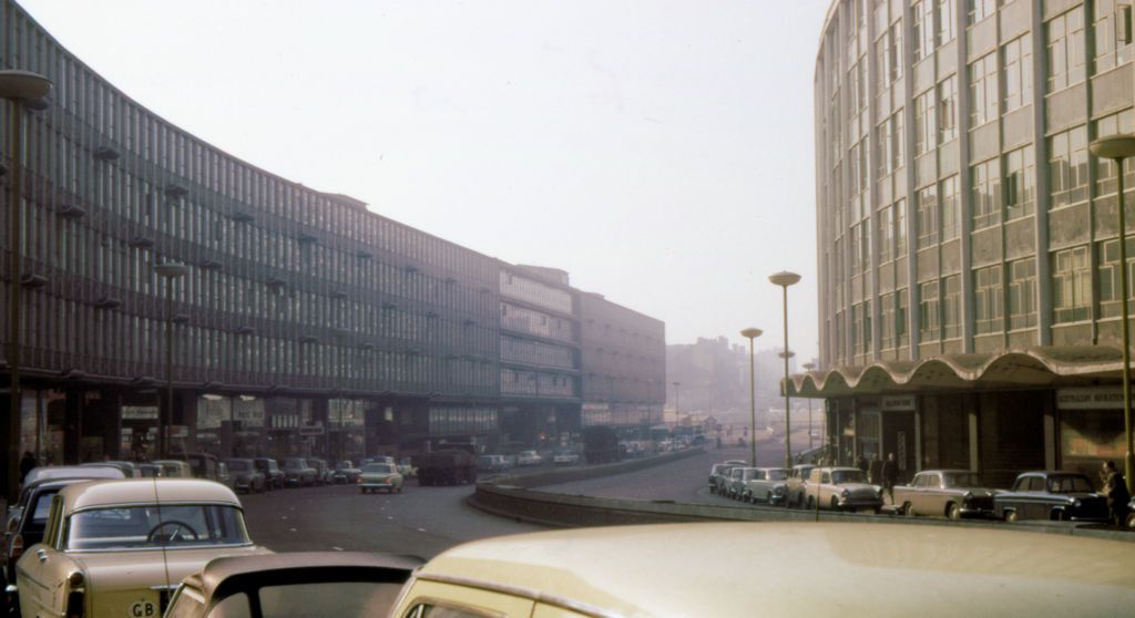 A long, curving block of shops and offices beside a sunken dual carriageway, with parked 1960s cars in the foreground and a tall building on the right.