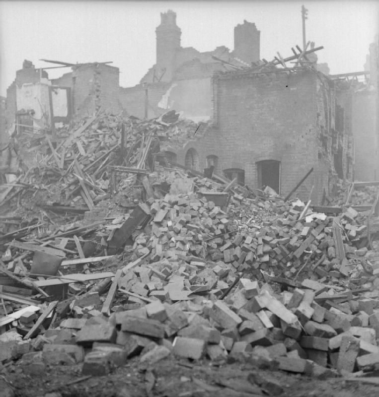 A heap of brick rubble where a terraced house stood, with damaged houses behind.