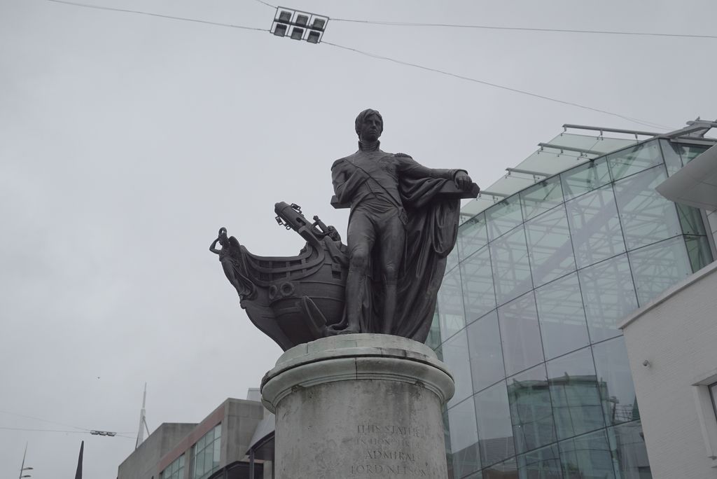 Bronze statue of Nelson beside a ship’s prow on a plain round stone pedestal, with a modern glass building behind.