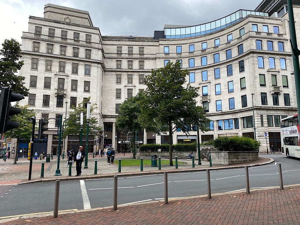 A small paved square with trees and bollards in front of a large curved Portland stone building.