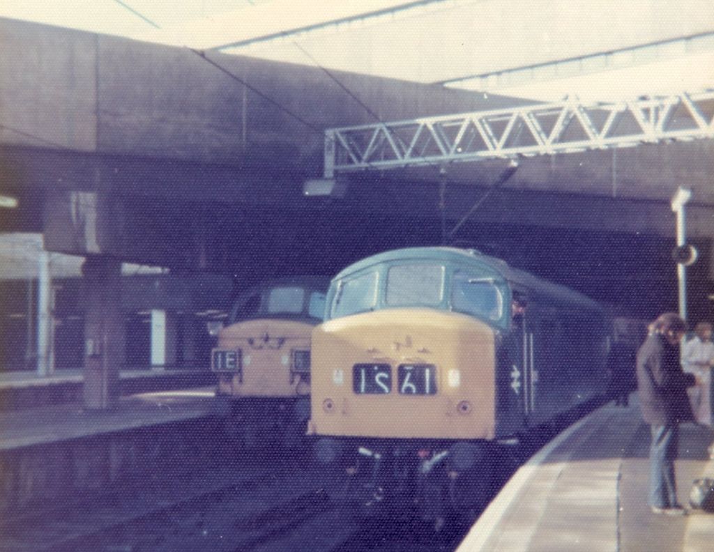 Colour photograph of two diesel locomotives at platforms beneath a low concrete deck.