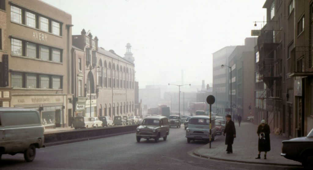 A busy street of shops and offices with cars and pedestrians, fading into haze in the distance.