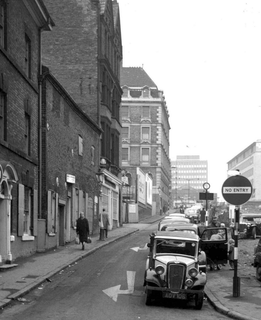 A narrow street of older buildings rising uphill, with a parked car and a No Entry sign.