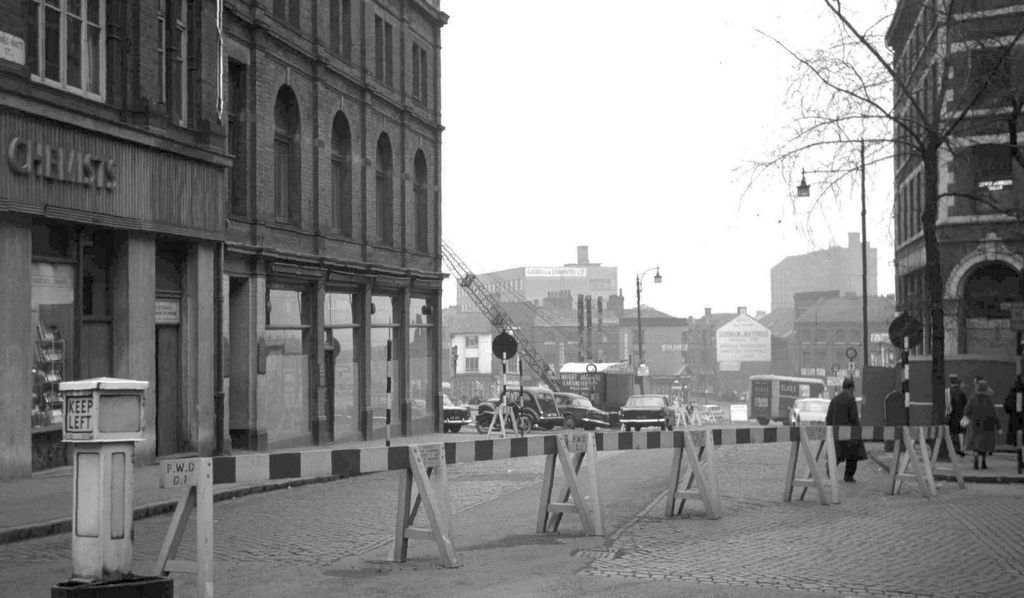 A city street with roadworks barriers, Victorian buildings on the left and cranes in the distance.