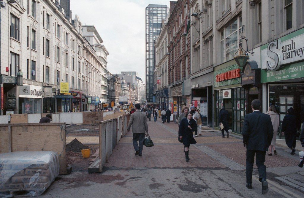 Shoppers walking along a city shopping street lined with Victorian buildings, with hoardings and street works on one side.