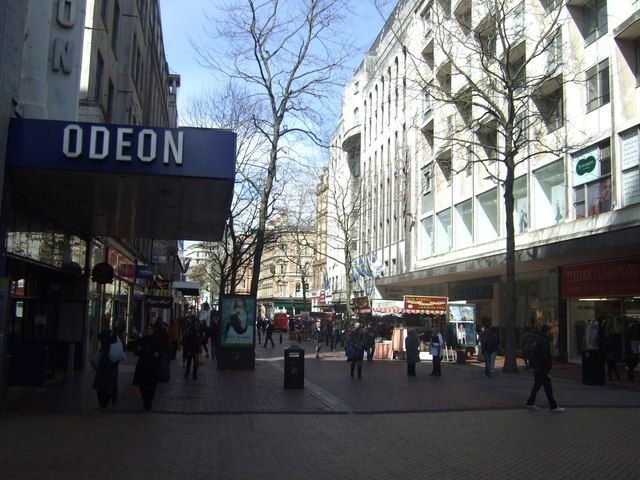 A paved pedestrian shopping street with trees, market stalls and shoppers, and a cinema sign on the left.