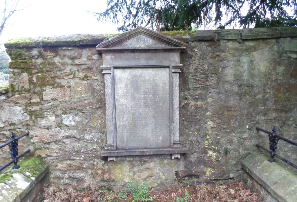 A weathered pedimented stone tablet set into an old rubble wall, flanked by low iron railings.