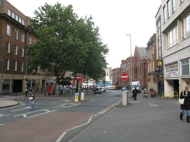 A city-centre street with a large tree, red-brick buildings on both sides, parked cars and people walking.