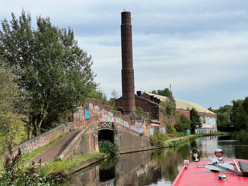 A tall brick chimney and old brick industrial buildings beside a canal, with a towpath bridge in front.