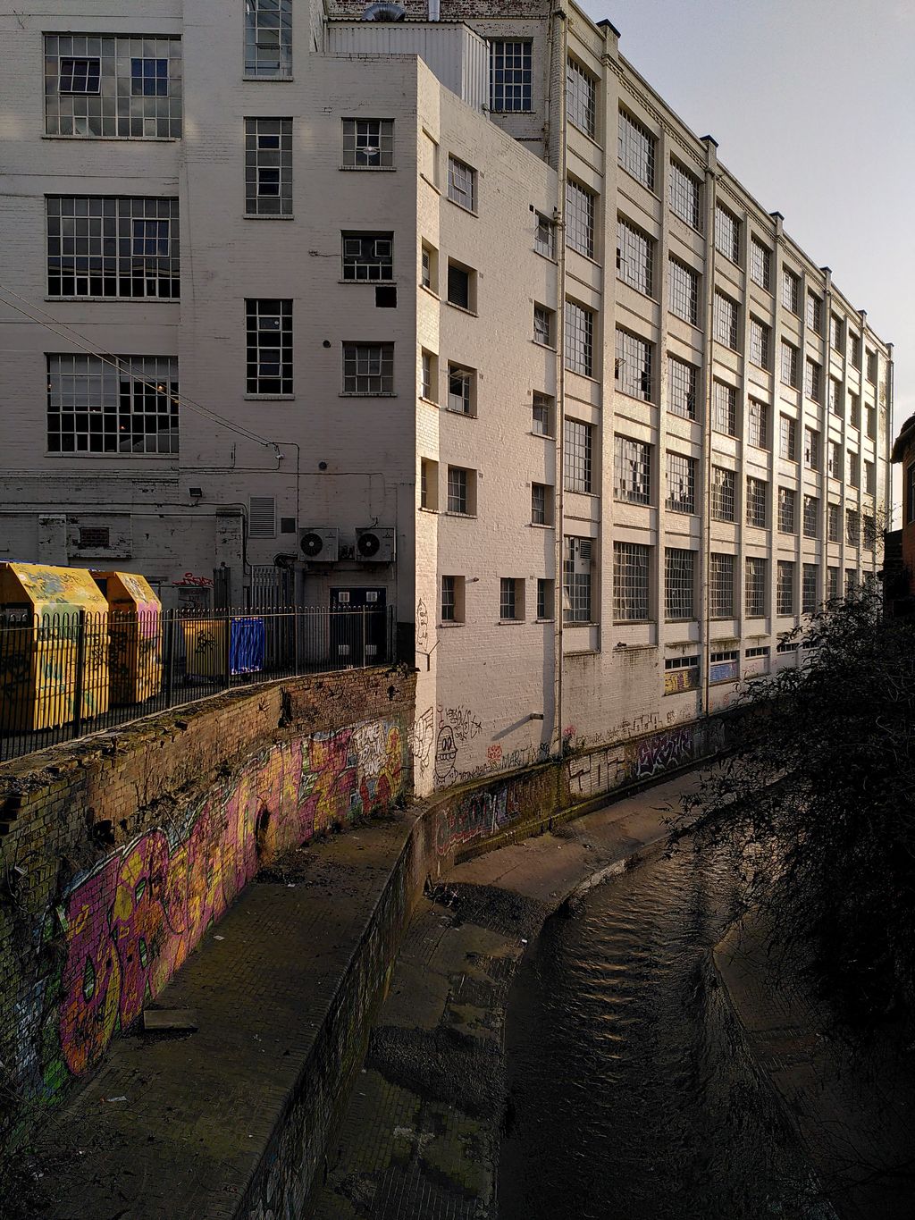 A narrow brick-lined river channel with graffiti, beside tall white-painted factory buildings.