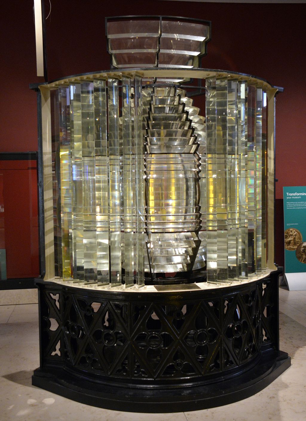 Tall glass lighthouse lens of stacked prisms in a brass frame, in a museum gallery.