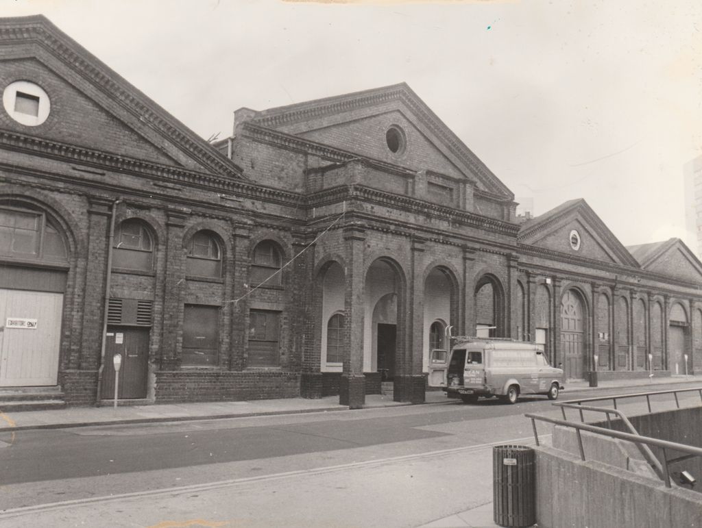 Black and white photograph of a brick building front with a pediment and round window above four open arches.