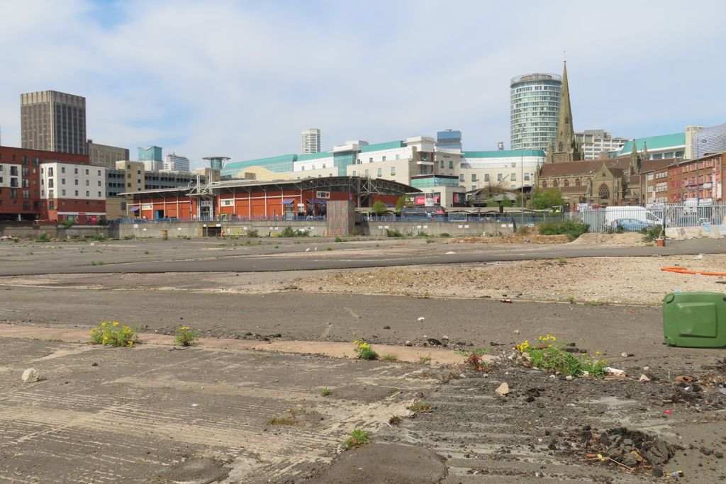 A wide cleared city-centre site behind fencing, with tower blocks beyond.