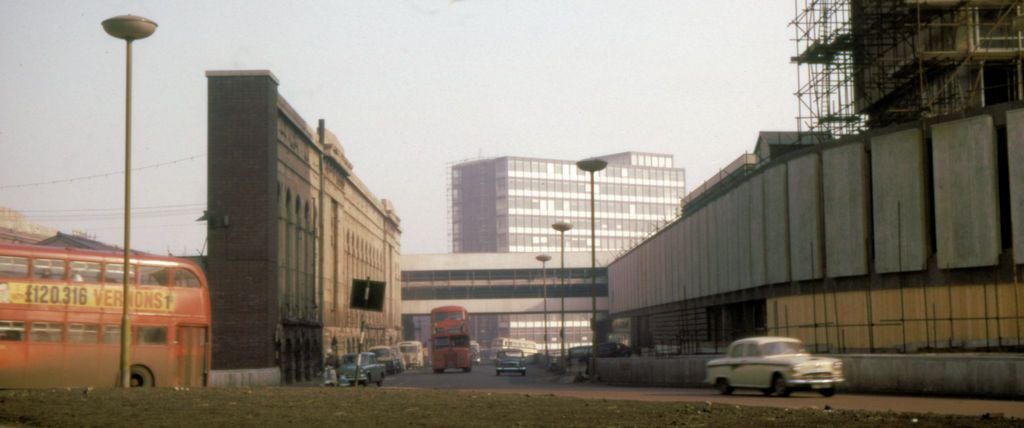 Black-and-white photograph of a road being built beside the side wall of a large old market hall, with hoardings on the other side.