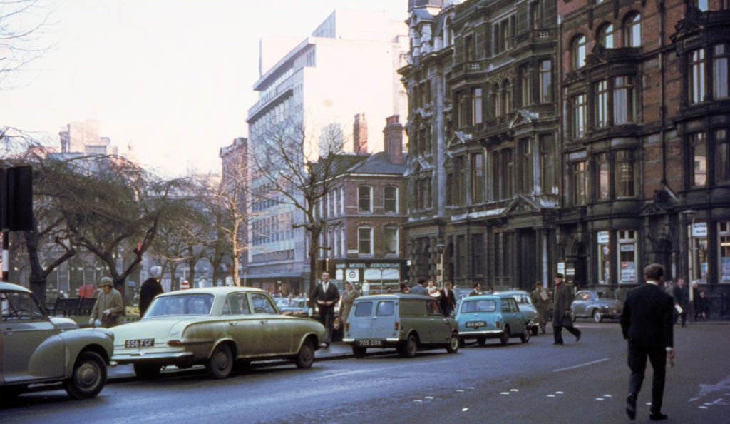 Black-and-white photograph of a city street with older shops and a new department store in the distance.
