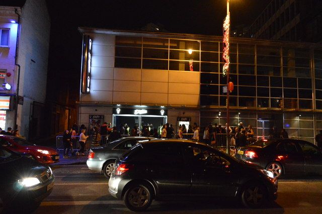 A modern glazed corner building lit up at night beside a city road.